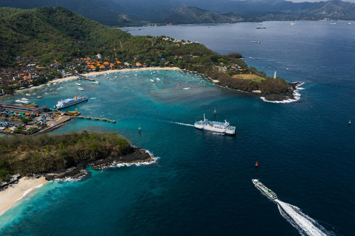 Aerial view of a coastal bay with a docked ferry and boats in clear turquoise waters, surrounded by green hills and a small coastal village with orange-roofed buildings.